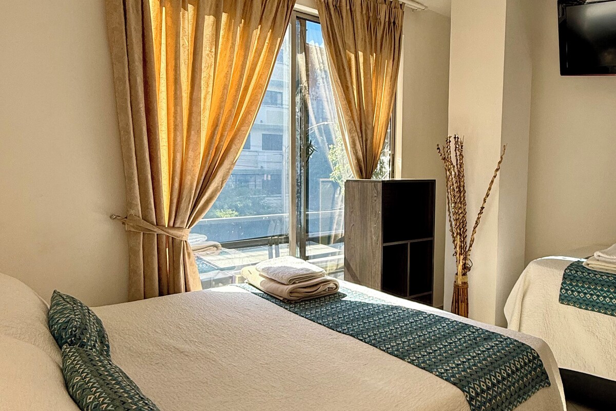 Ein Hotelzimmer mit einem großen Bett, einem kleineren Bett, einem Flachbildfernseher und Blick auf die Stadt durch große Fenster.