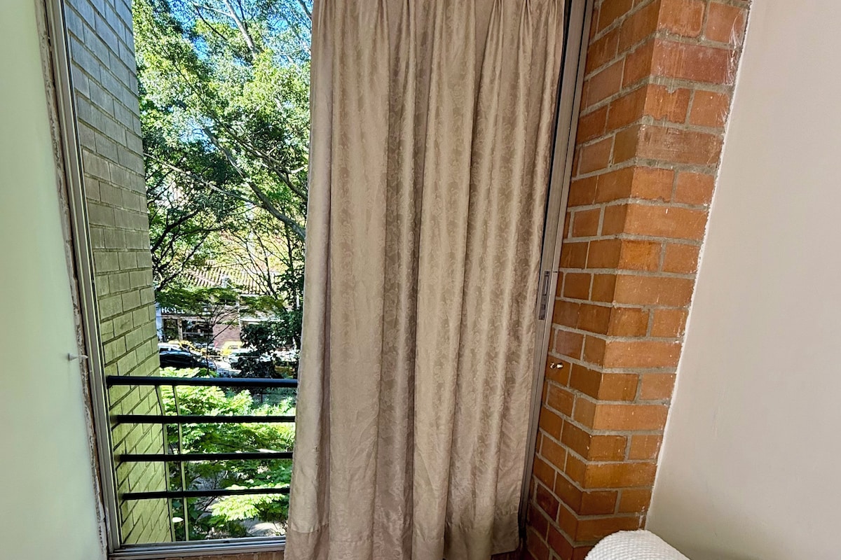Habitació amb llit, finestra amb cortines i vista a vegetació, juntament amb una paret de maó.