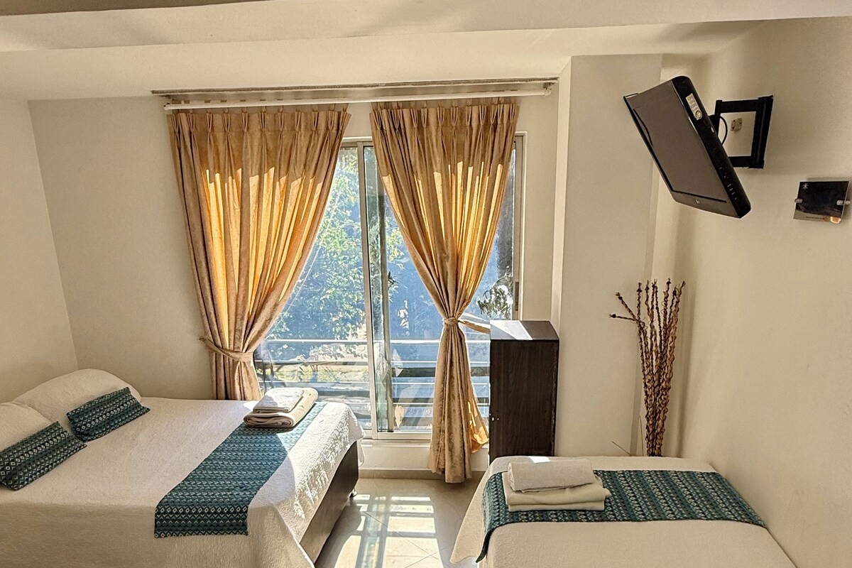 Habitación de hotel con dos camas, un ventanal con cortinas, un televisor de pantalla plana montado en la pared y una mesita.