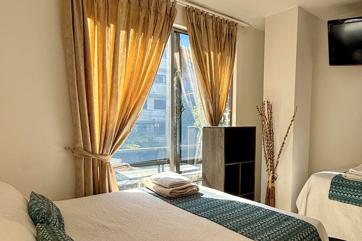 Une chambre d’hôtel avec un grand lit, un lit plus petit, un téléviseur à écran plat et une vue sur la ville grâce à de grandes fenêtres.