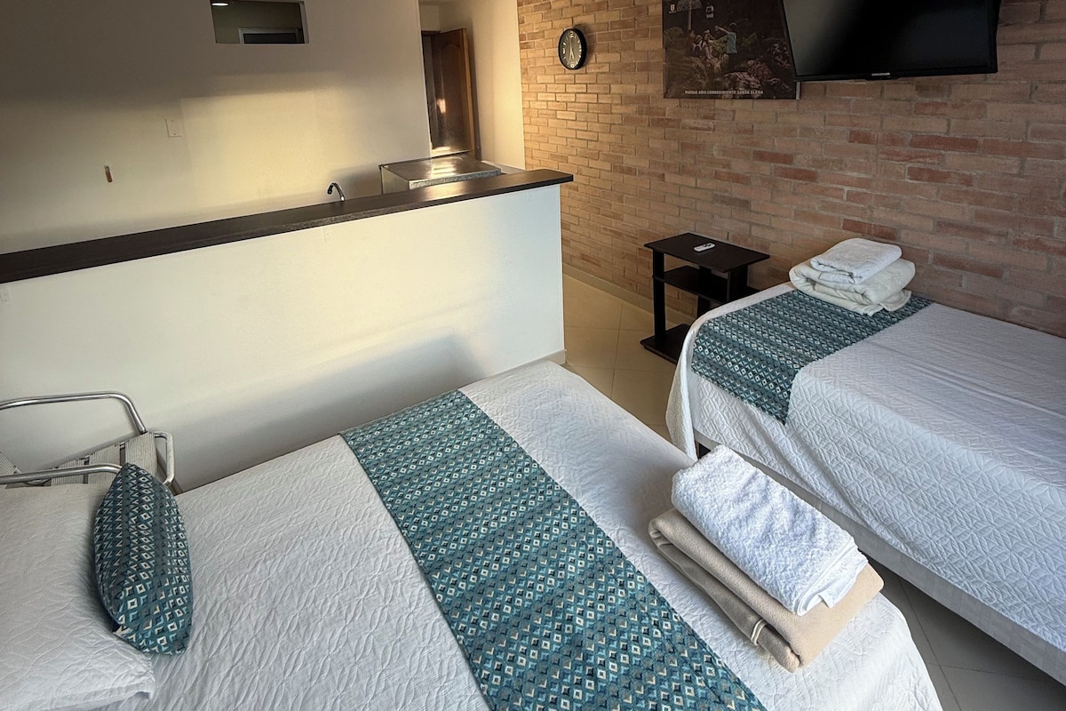 A hotel room with two beds, a TV mounted on a brick wall, a small table, and a clock.