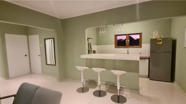 A modern kitchen with a white countertop, three bar stools, a refrigerator, and a window with a view.