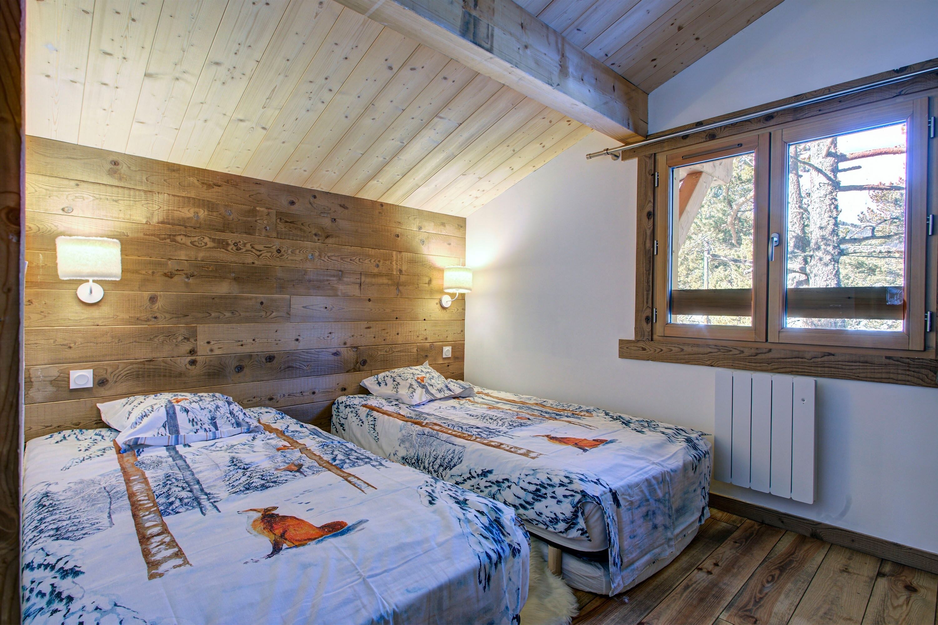 A room with two beds, wooden walls, a window with a view, and a wall-mounted light fixture.