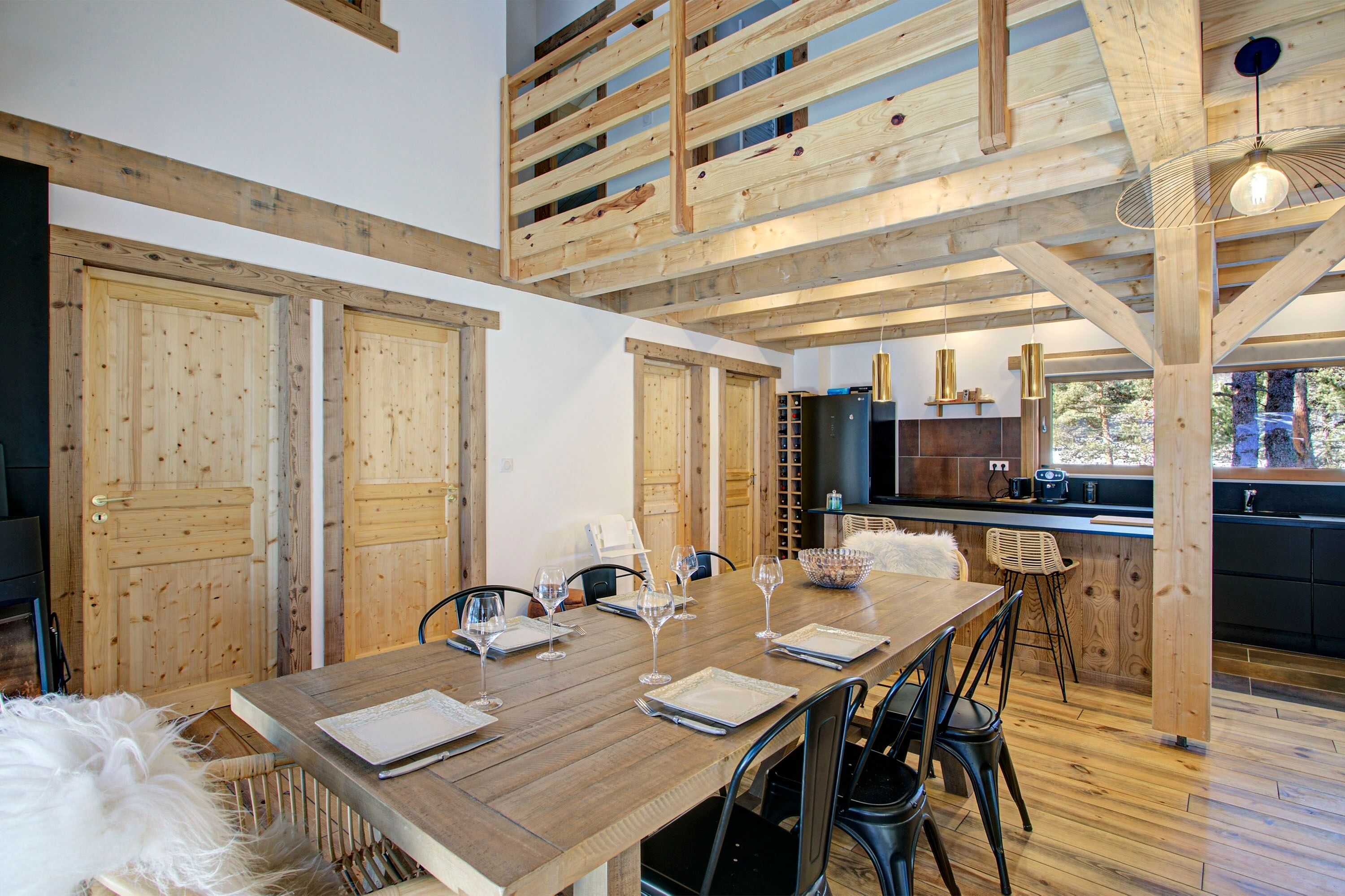 Una mesa de comedor de madera dispuesta para seis personas, copas de vino y platos, una cocina con electrodomésticos modernos y un altillo con vigas de madera.