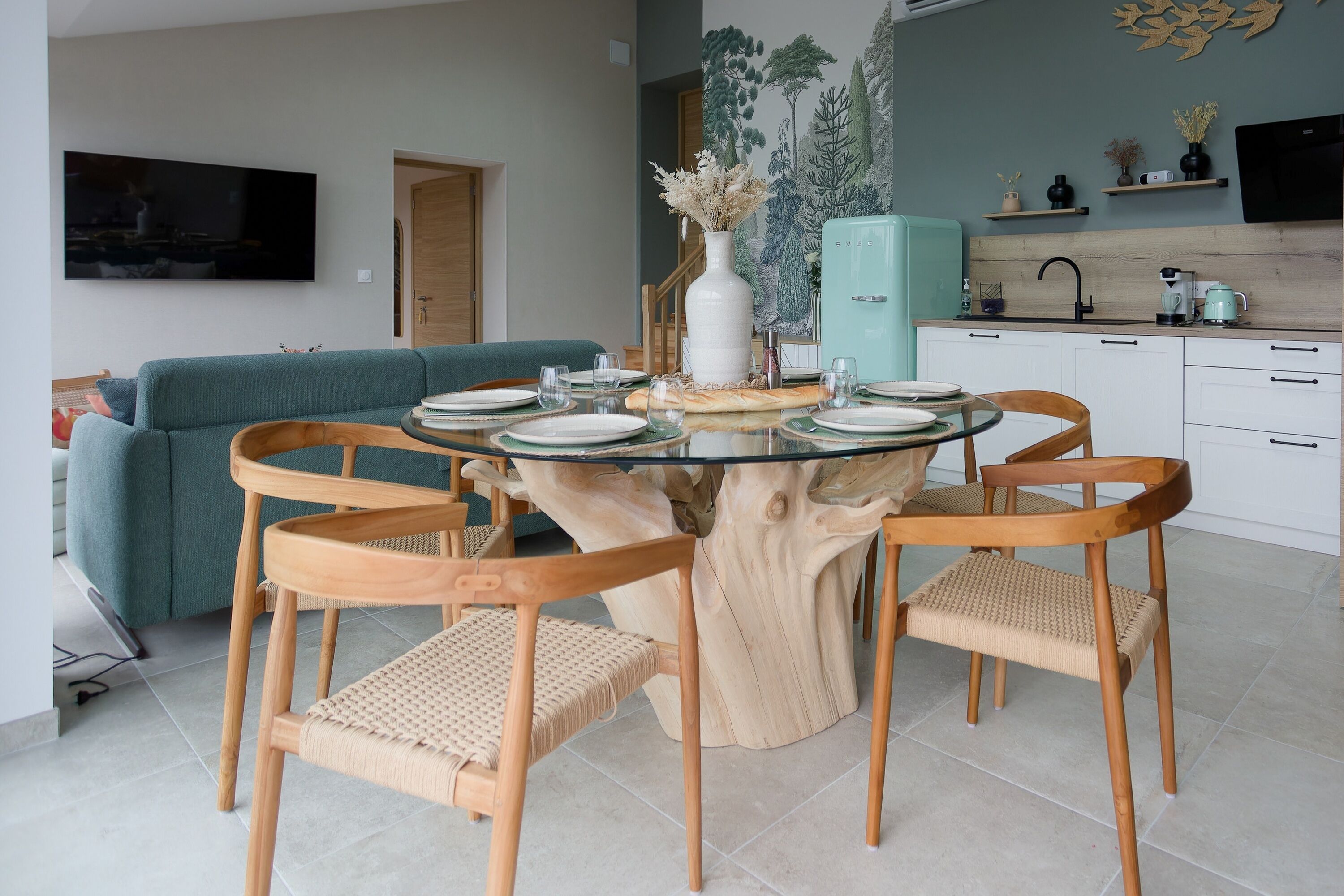 Un coin repas avec une table ronde en verre entourée de chaises en bois, un canapé de couleur turquoise et un espace cuisine avec un réfrigérateur turquoise et des placards en bois.