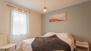 Une chambre à coucher avec un lit, une table de chevet, une chaise, une fenêtre avec des rideaux et un tableau au mur.