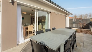 Une terrasse avec une table et des chaises, une porte-fenêtre coulissante donnant sur une salle à manger intérieure, et une cour arrière clôturée.
