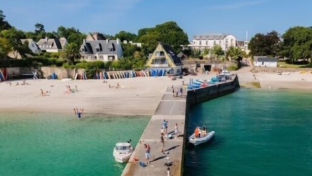 An aerial view of a beach with a pier extending into the water, people walking along the pier, and boats docked nearby.