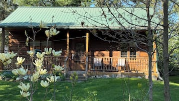 A wooden cabin with a green roof, a porch with a railing, and magnolia trees in the foreground.