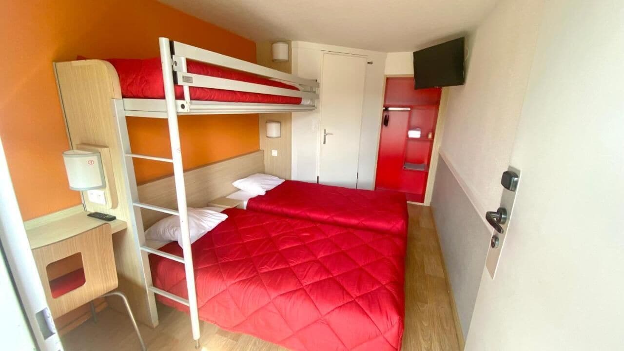 A bunk bed room with a red bedspread, a desk with a chair, a television, and a red wardrobe.