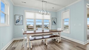 A dining room with a wooden table and benches, large windows with views, and two wall-mounted bird paintings. - Simply Blessed- 5BR/5.5BA Channel House-Pool (North Myrtle Beach)