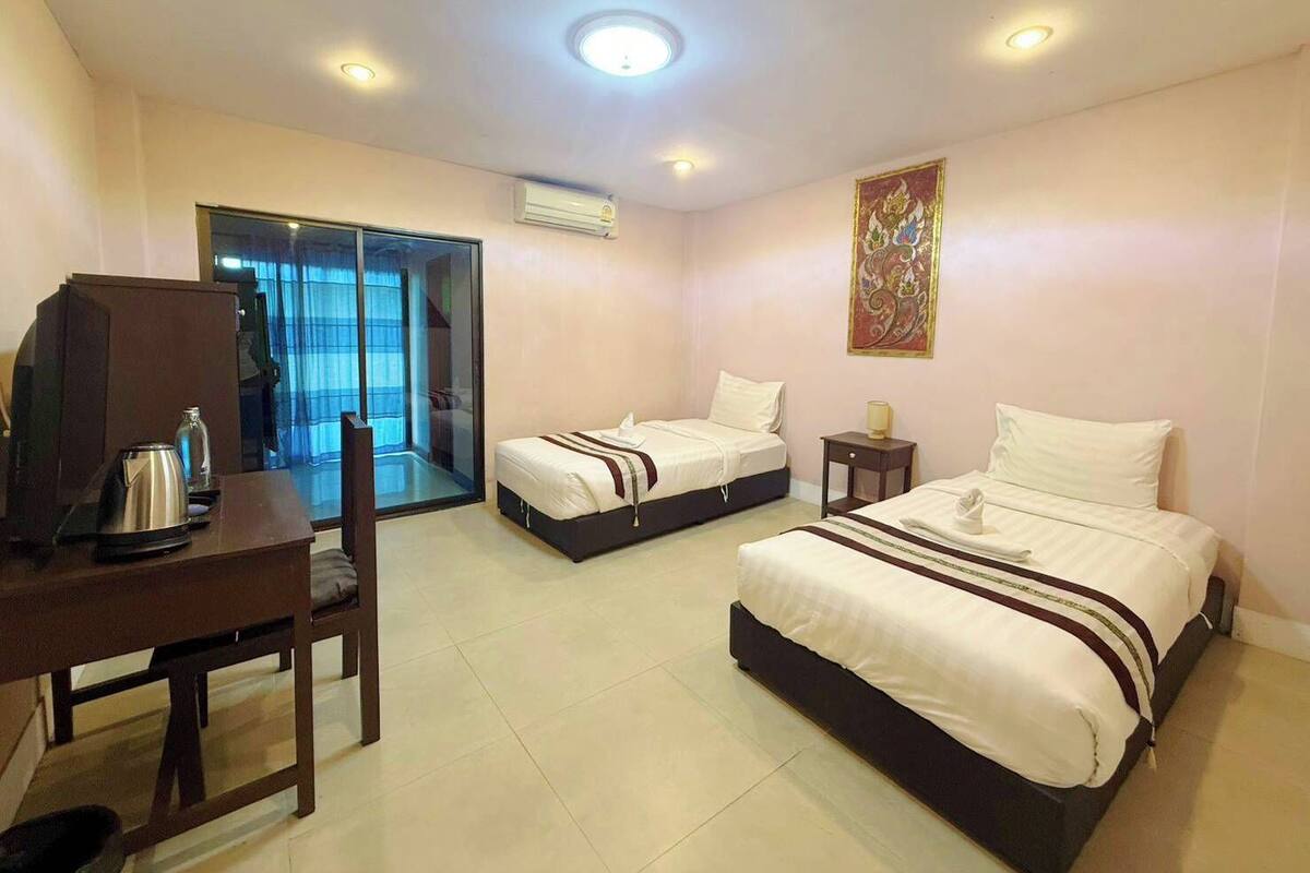 A hotel room with two beds, a desk with a kettle, a small table, a chair, a sliding glass door, and a ceiling light.