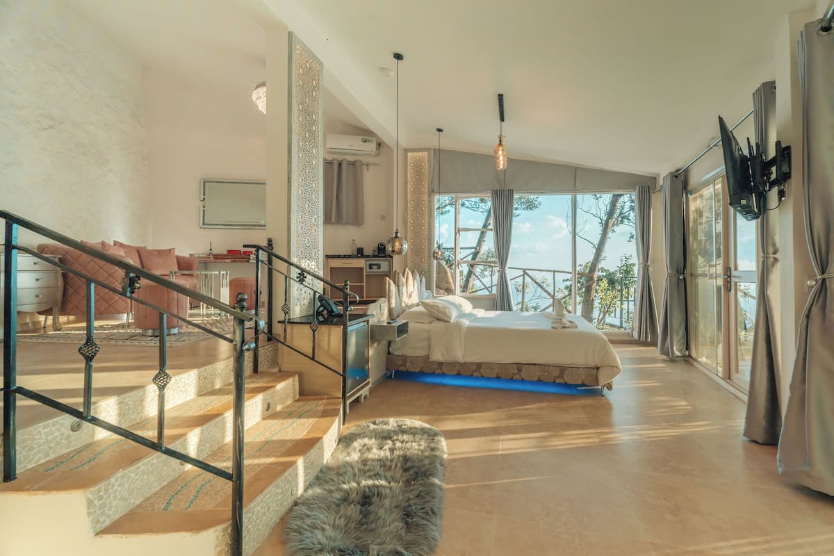 Une chambre spacieuse avec un grand lit, un escalier doté d’une rampe en métal et une vue sur l’extérieur grâce à de grandes fenêtres.