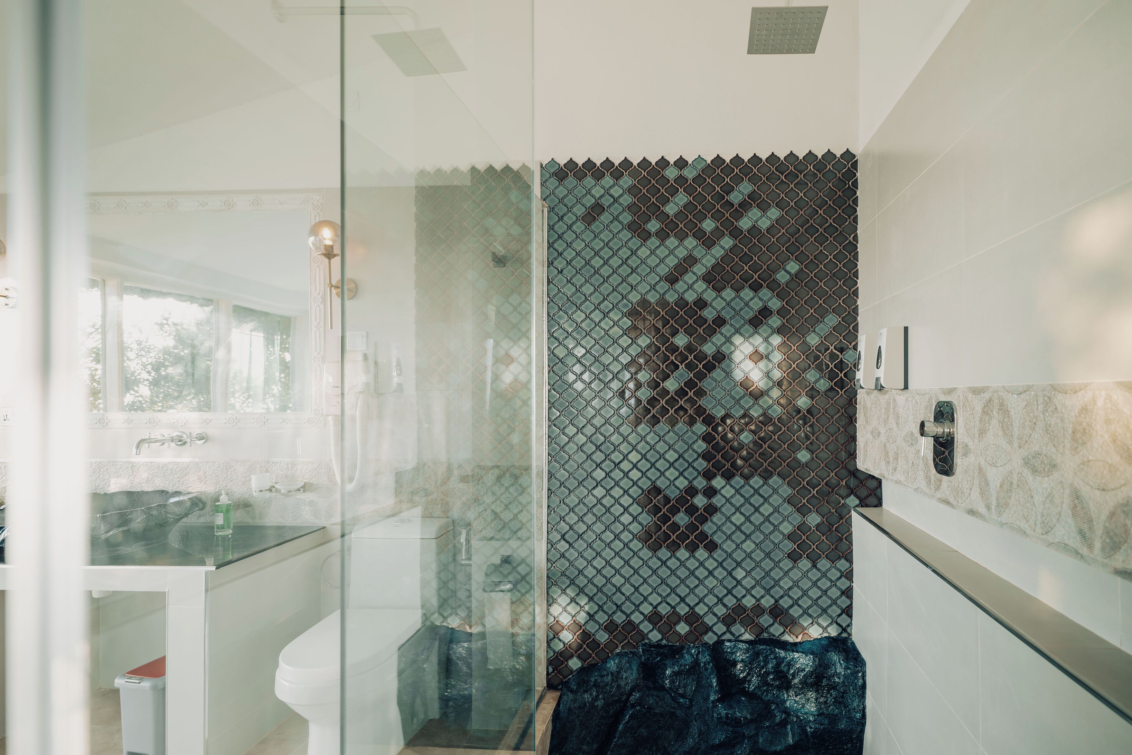 Une salle de bain moderne avec une douche à l’italienne en verre, un lavabo blanc et un mur recouvert de carreaux à motifs.
