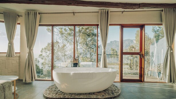 A spacious bathroom with a large freestanding bathtub, large windows with curtains, and a view of the outdoors.