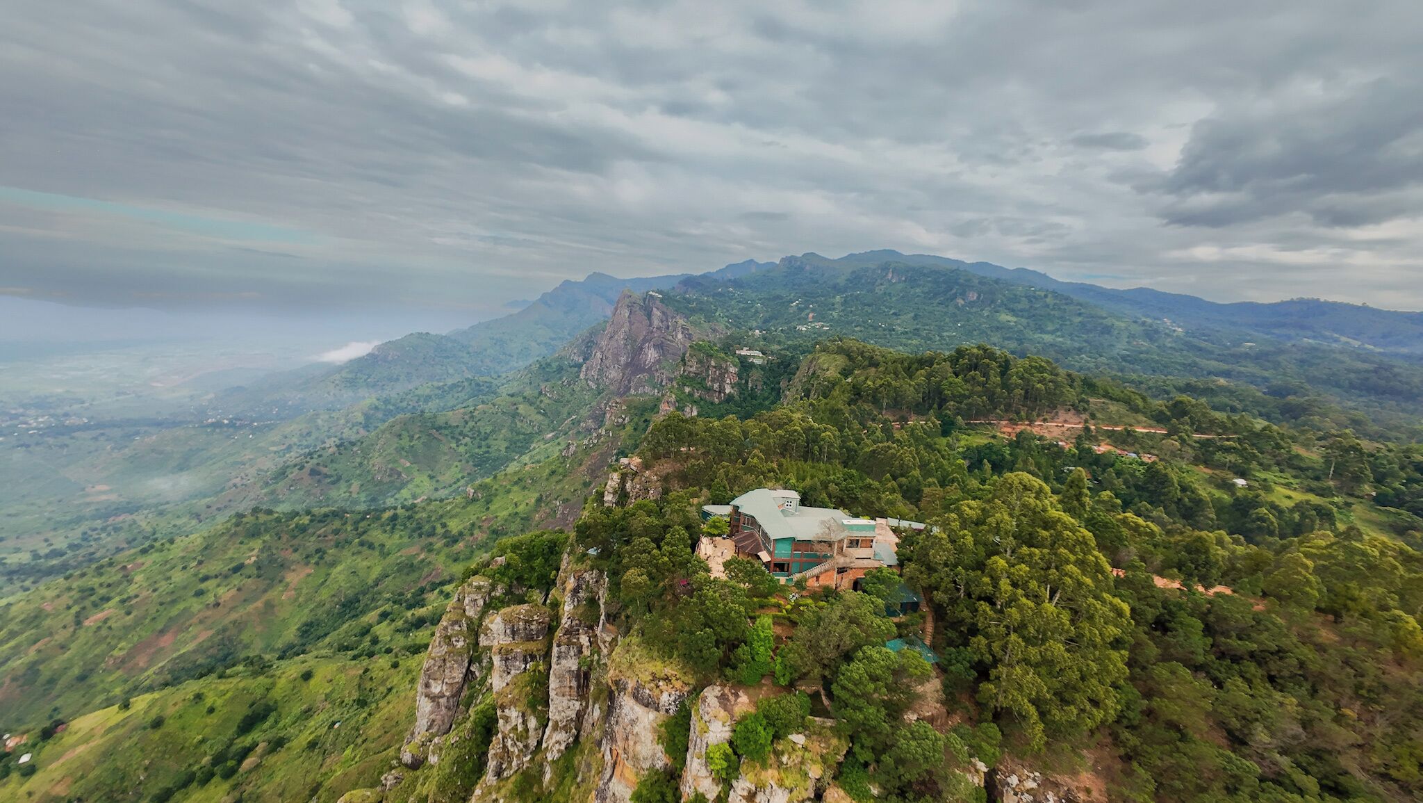 A mountain house with a green roof, surrounded by lush greenery and rocky cliffs.