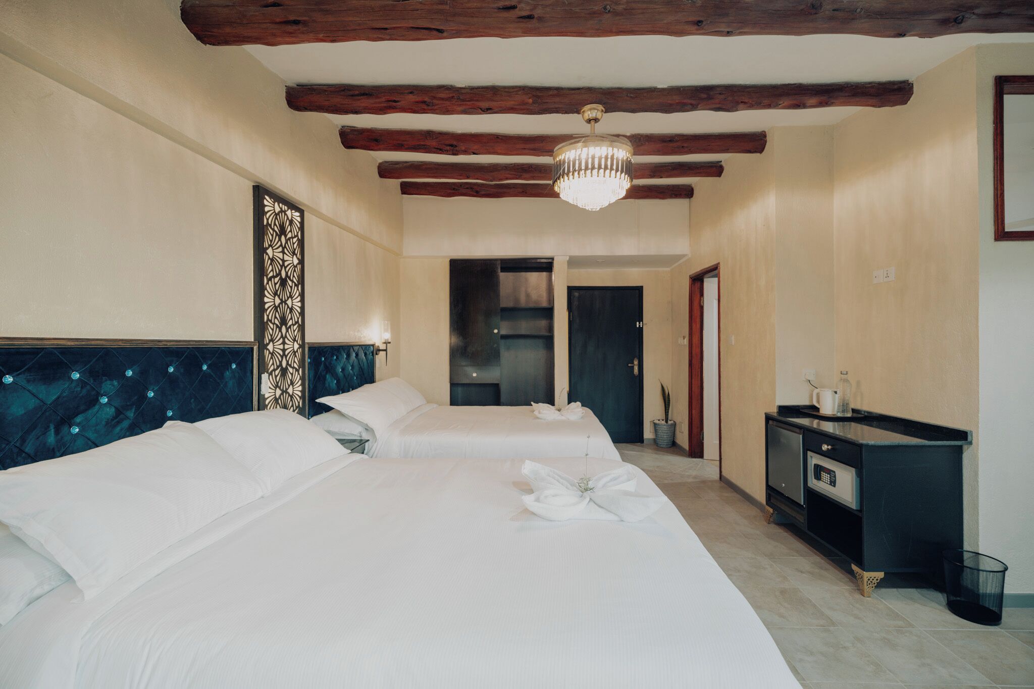 A hotel room with two beds, a chandelier, a minibar, and a door leading to another room.