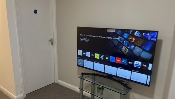 A flat-screen TV mounted on a wall with a glass stand below it. - 5 Bedroom Cosy Bloxwich Stay - Wifi & Free Parking (Walsall)