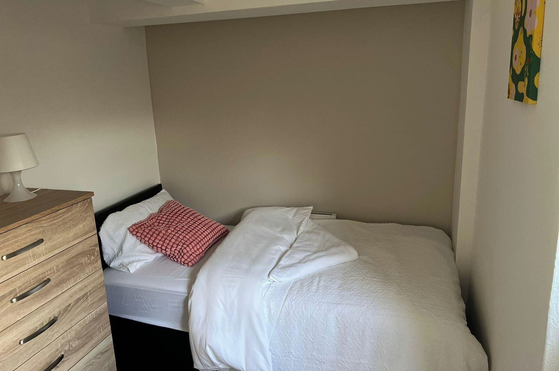 Un petit lit bien fait, avec une literie blanche et un coussin à motifs rouges, dans une pièce simple comprenant une commode en bois et une lampe.
