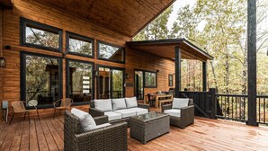 Une terrasse en bois, meublée de meubles en osier, dotée de grandes fenêtres et offrant une vue sur les arbres.