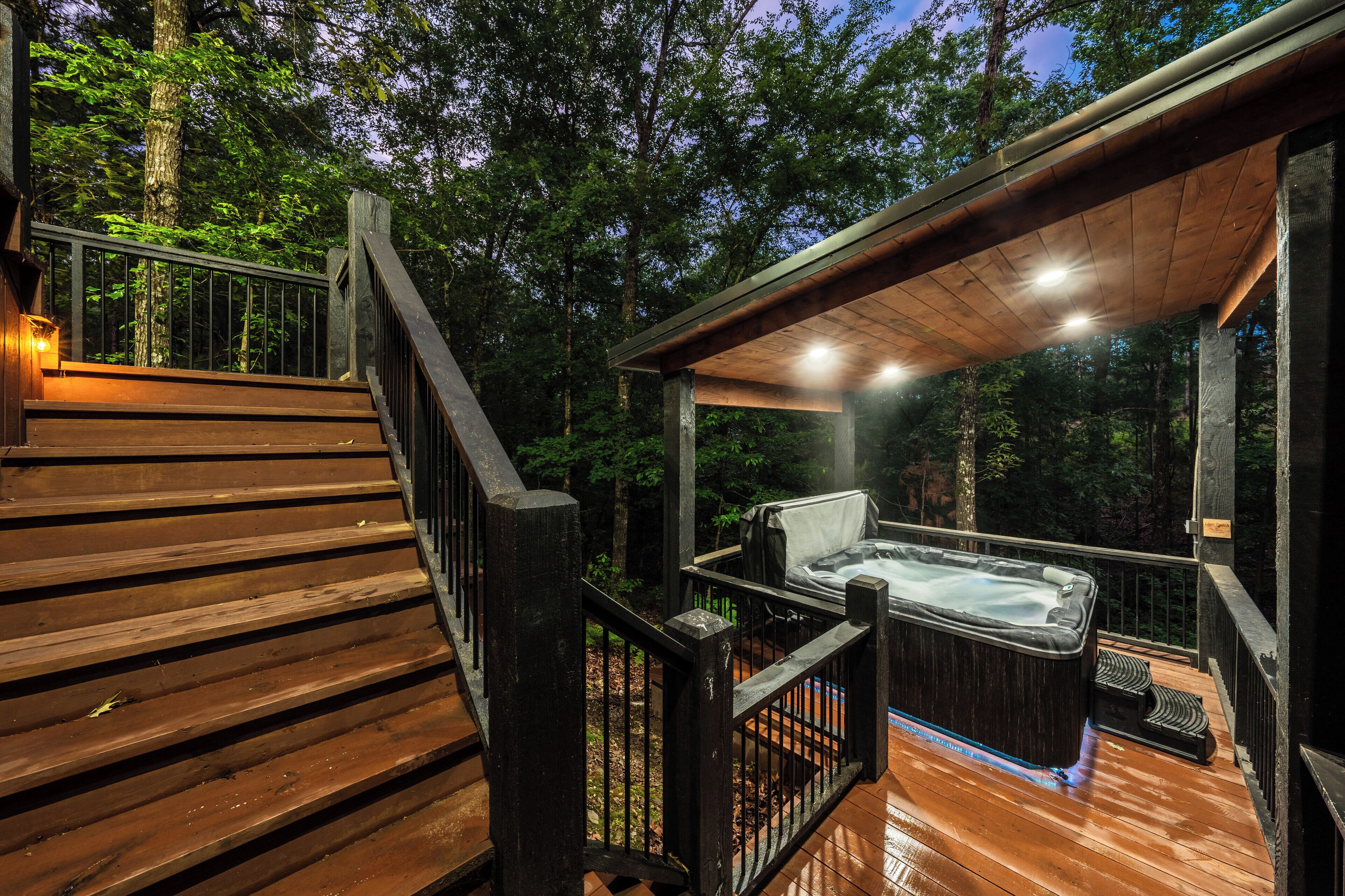A wooden deck with a hot tub and a covered seating area, surrounded by trees.