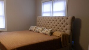 Ein Schlafzimmer mit einem großen Bett, einem gesteppten Kopfteil und zwei Fenstern mit Jalousien.