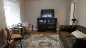 Ein Wohnzimmer mit einer Couch, einem Sessel, einem Fernseher auf einem Schrank und einem Fenster mit Jalousien.