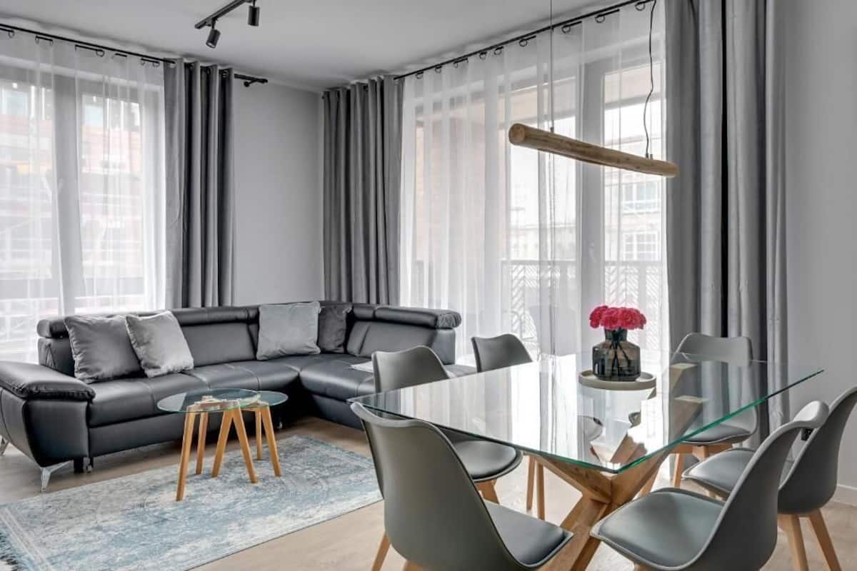 A modern living room with a glass dining table, grey sofa, and a large window with sheer curtains.