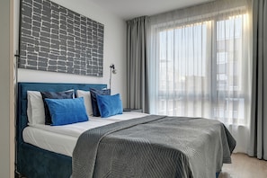 Habitación de hotel moderna con una cama grande, un cabecero azul y vistas de la ciudad a través de cortinas translúcidas.