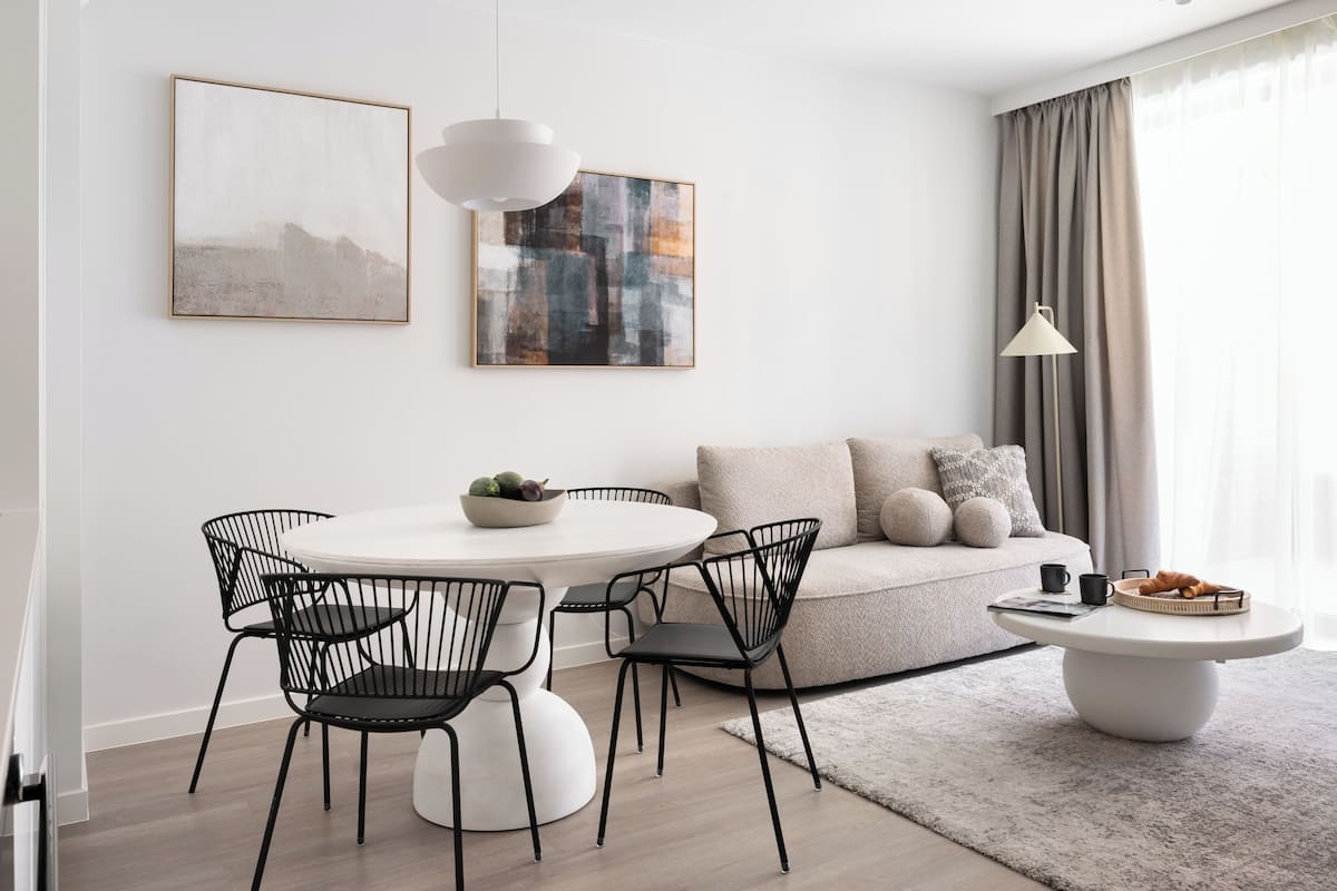 A modern living room with a round dining table, black wire chairs, a white coffee table, and a beige sofa with cushions. There are abstract paintings on the wall and a floor lamp.
