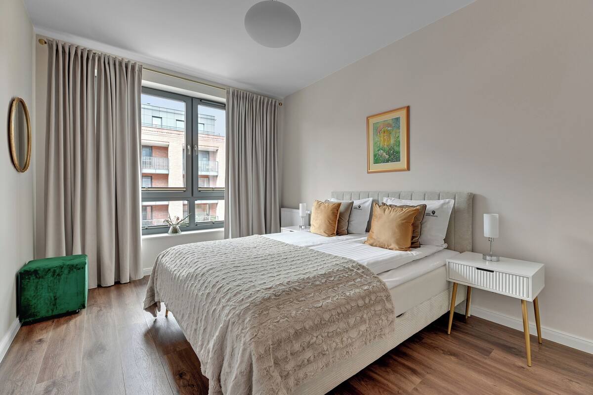A bedroom with a large bed, a bedside table, a green ottoman, and a window with curtains.