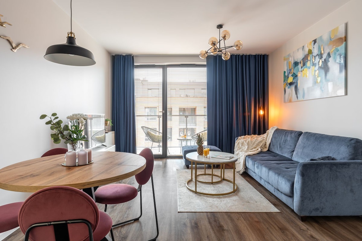 A modern living room with a blue sofa, a round wooden dining table with chairs, a small coffee table, and a large window with curtains.