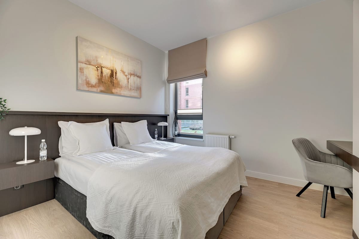 A modern hotel room with a large bed, a desk, and a chair. There is a window with a view of buildings outside.