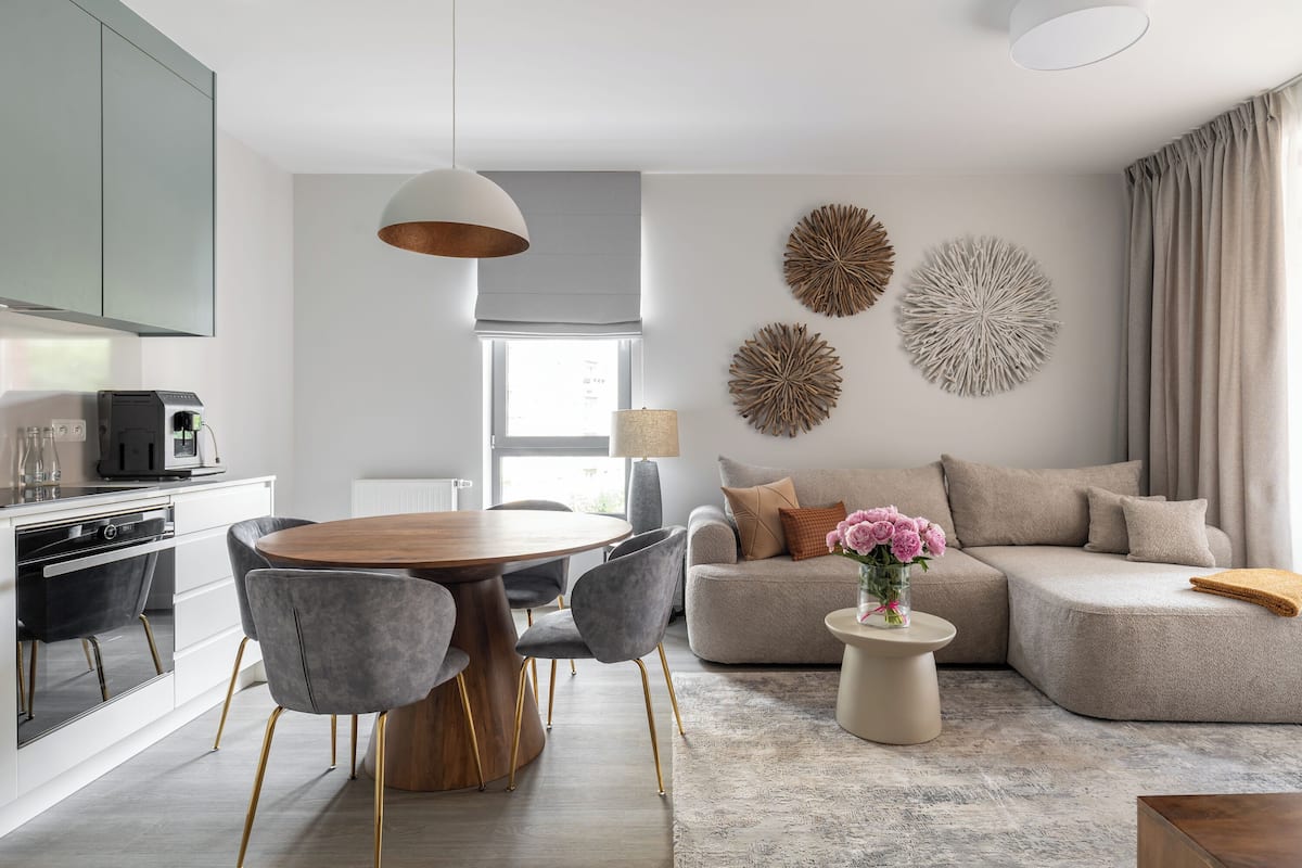A modern living room with a round wooden dining table, grey upholstered chairs, a beige sofa, and decorative wall art.