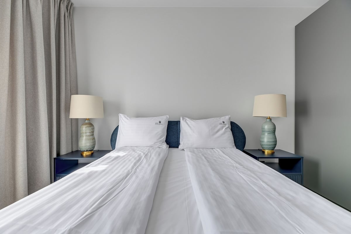 A double bed with white linens, two bedside tables with lamps, and a curtain on the left.