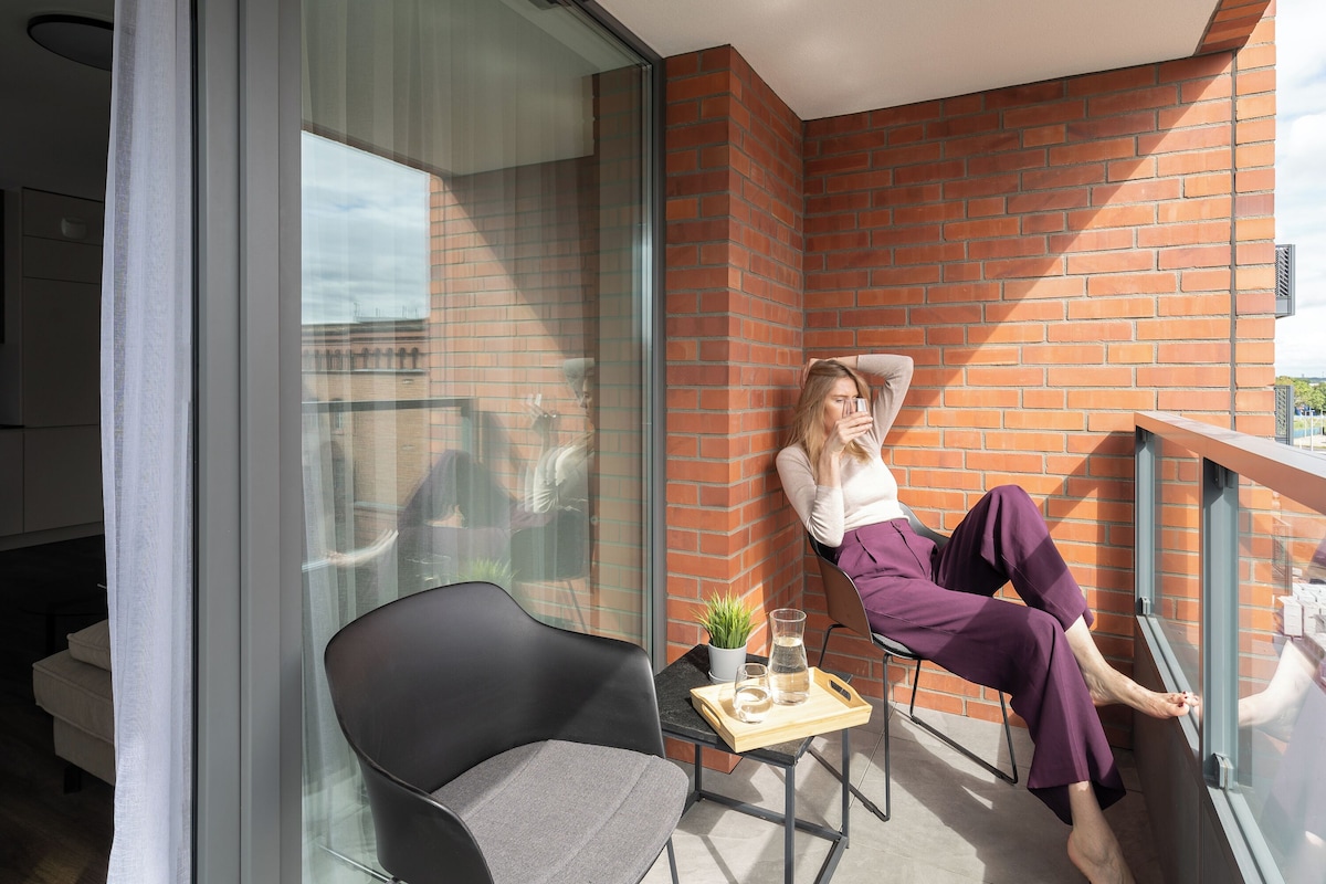A balcony with a modern design, featuring a glass partition, a black chair, and a small table with a plant and a glass.