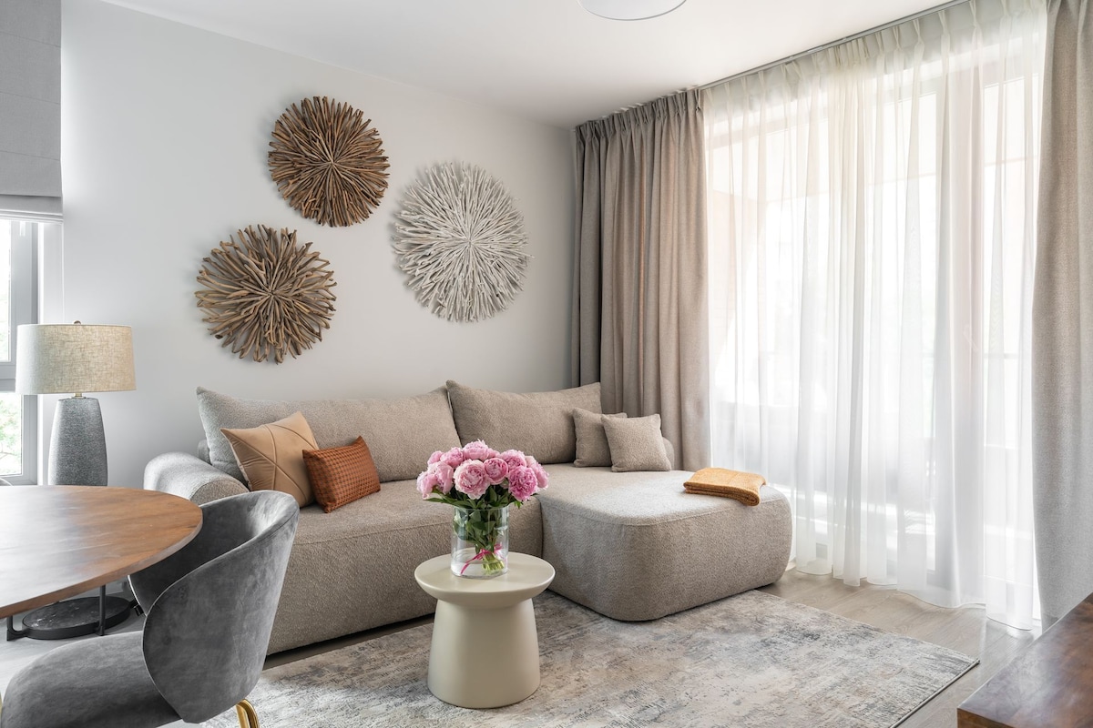 A modern living room with a sofa, ottoman, and a round coffee table with a vase of flowers.