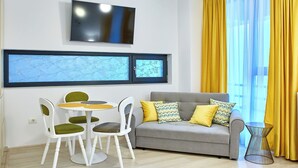 Una sala de estar moderna con un sofá gris, cojines amarillos, una mesa de comedor blanca con sillas verdes y un televisor montado en la pared.