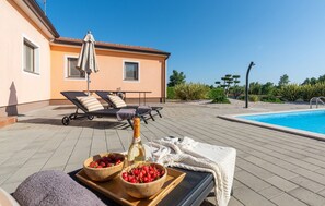 Un espace aménagé au bord de la piscine, avec des chaises longues, une table sur laquelle sont disposés des fruits et une bouteille, et un bâtiment en arrière-plan.
