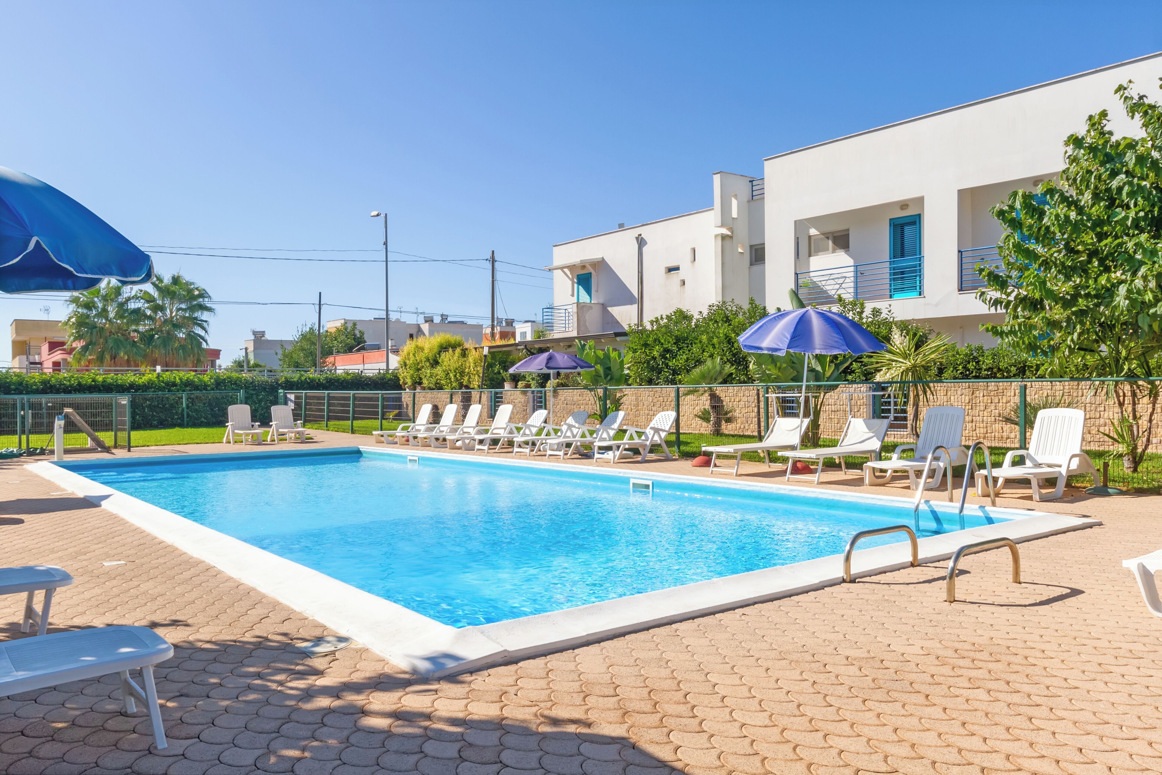 Una piscina rectangular con agua azul cristalina, rodeada de sillas reclinables blancas y sombrillas azules.