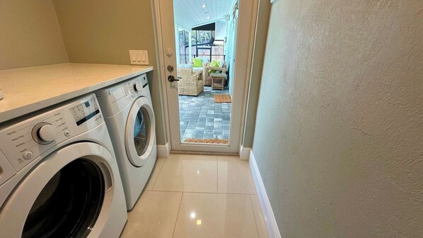 Une buanderie équipée d’un lave-linge et d’un sèche-linge, avec un sol carrelé et une porte donnant sur un espace de vie comprenant un canapé et une table basse.