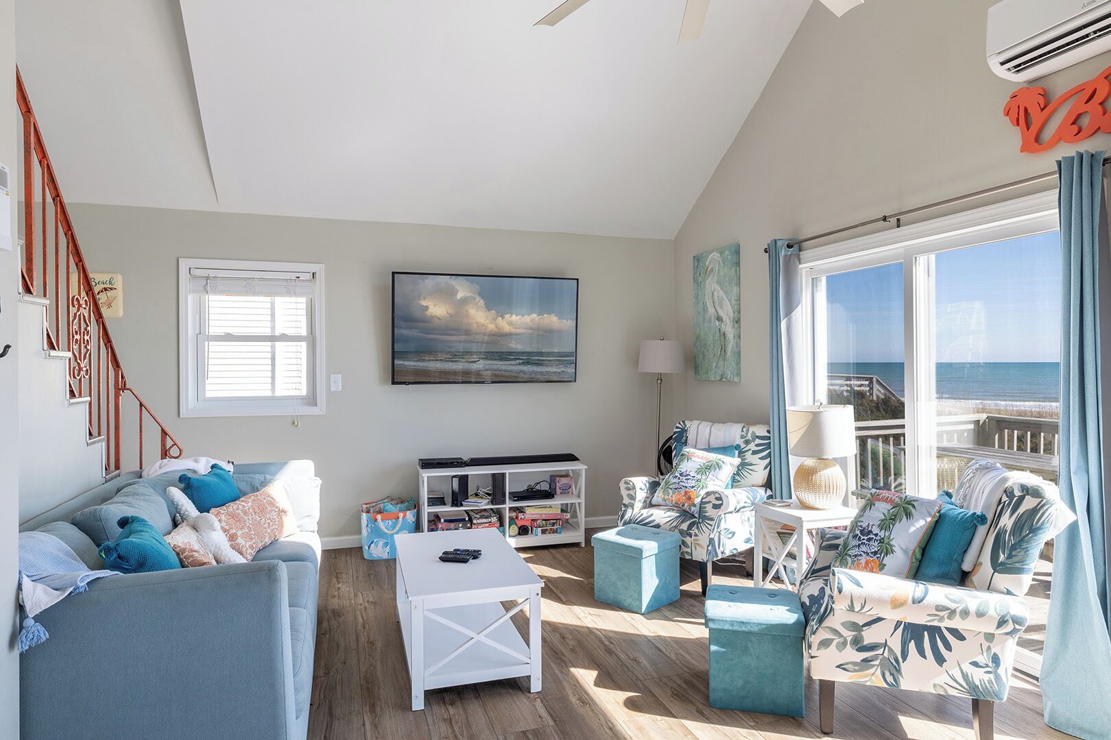 Un salón amplio con un gran ventanal con vistas a la playa, una escalera y un cuadro colgado.