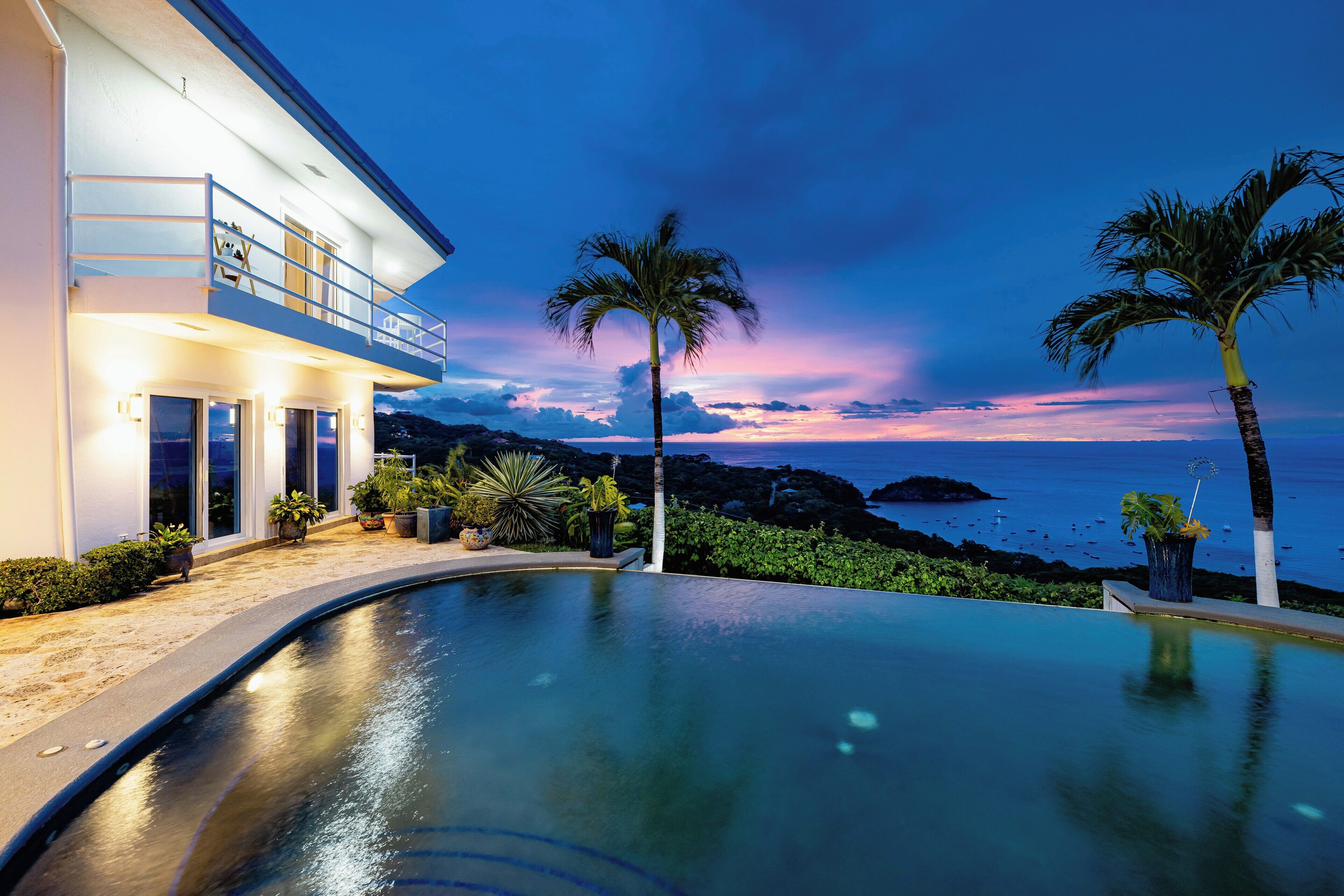Uma casa moderna com piscina, varanda e vista para o oceano ao entardecer.