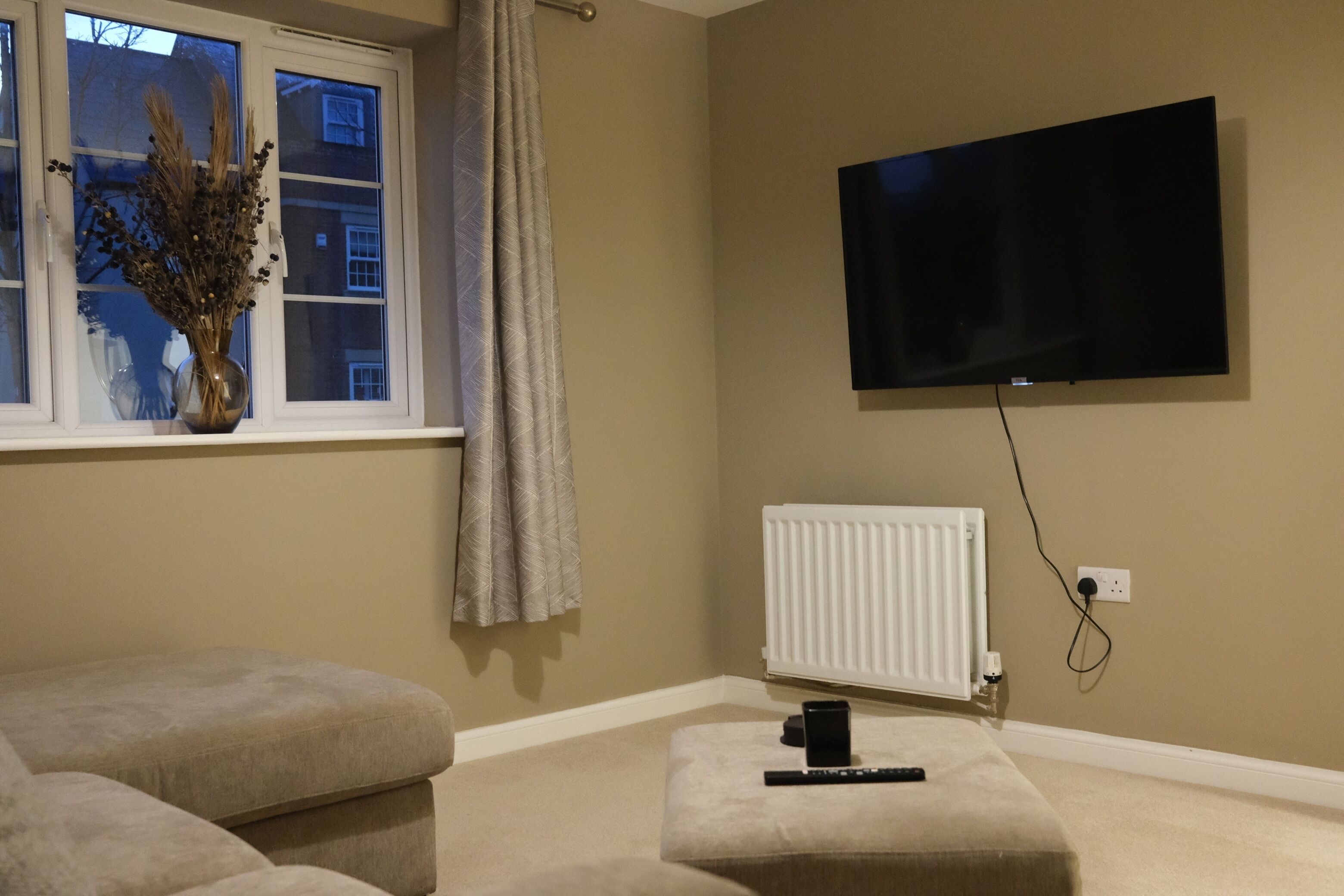 Een woonkamer met een flatscreen televisie aan de muur, een radiator eronder en een raam met gordijnen.