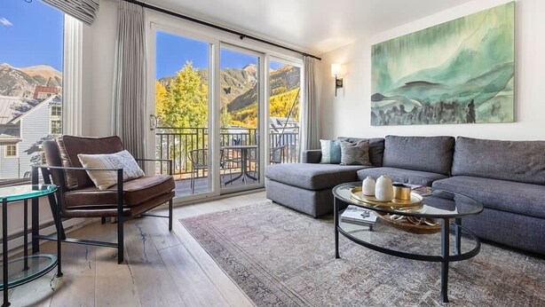 A modern living room with a sofa, armchair, and coffee table, large sliding glass doors leading to a balcony with a mountain view.