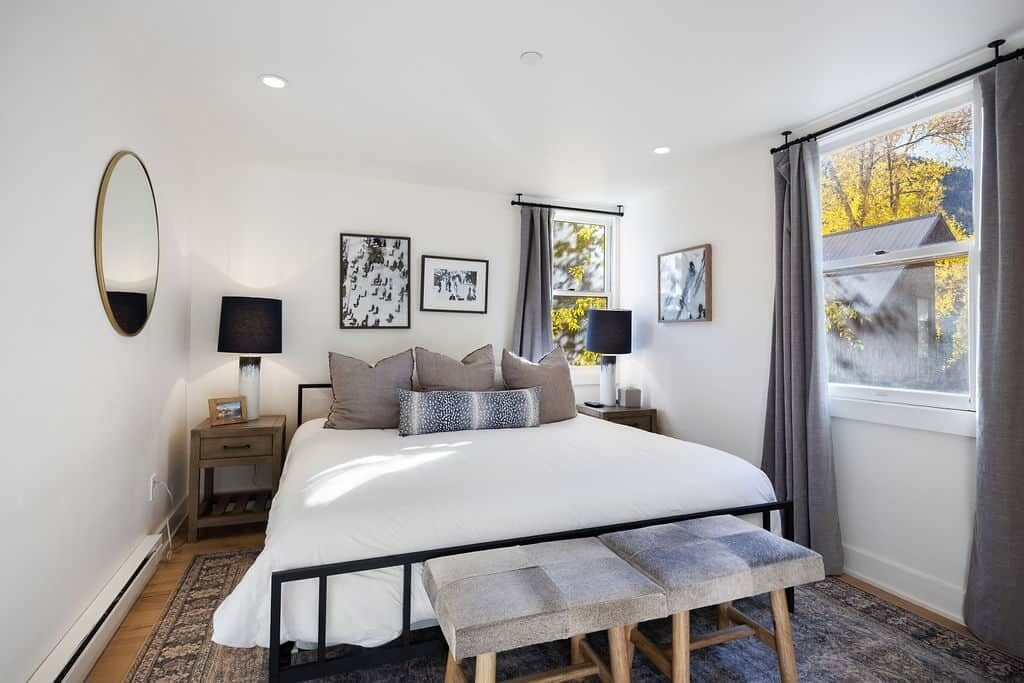 Un dormitorio con cama, mesitas de noche, lámparas, cuadros enmarcados, una ventana con cortinas y vista a árboles.