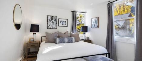 Un dormitorio con cama, mesitas de noche, lámparas, cuadros enmarcados, una ventana con cortinas y vista a árboles.