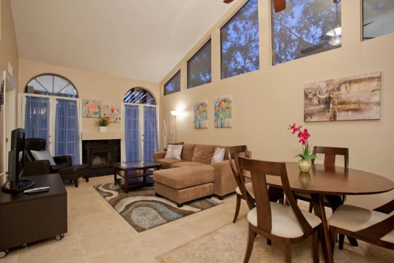 A spacious living room with a fireplace, a sofa set, a dining table with chairs, and large windows.