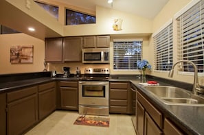 A modern kitchen with stainless steel appliances, a large window with blinds, and a decorative rug on the floor.
