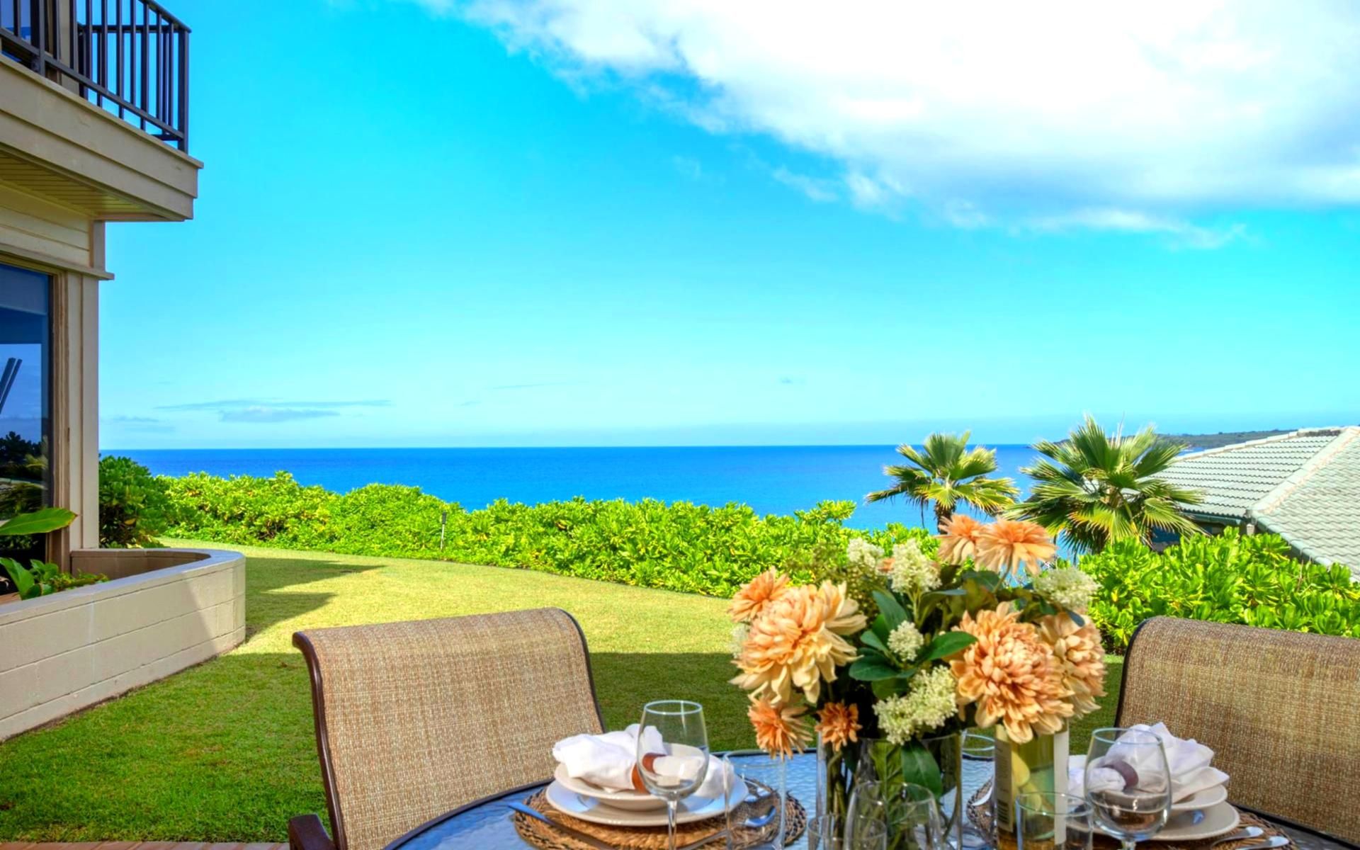 An outdoor dining area with a view of the ocean, a table set with plates, glasses, and a vase of flowers.